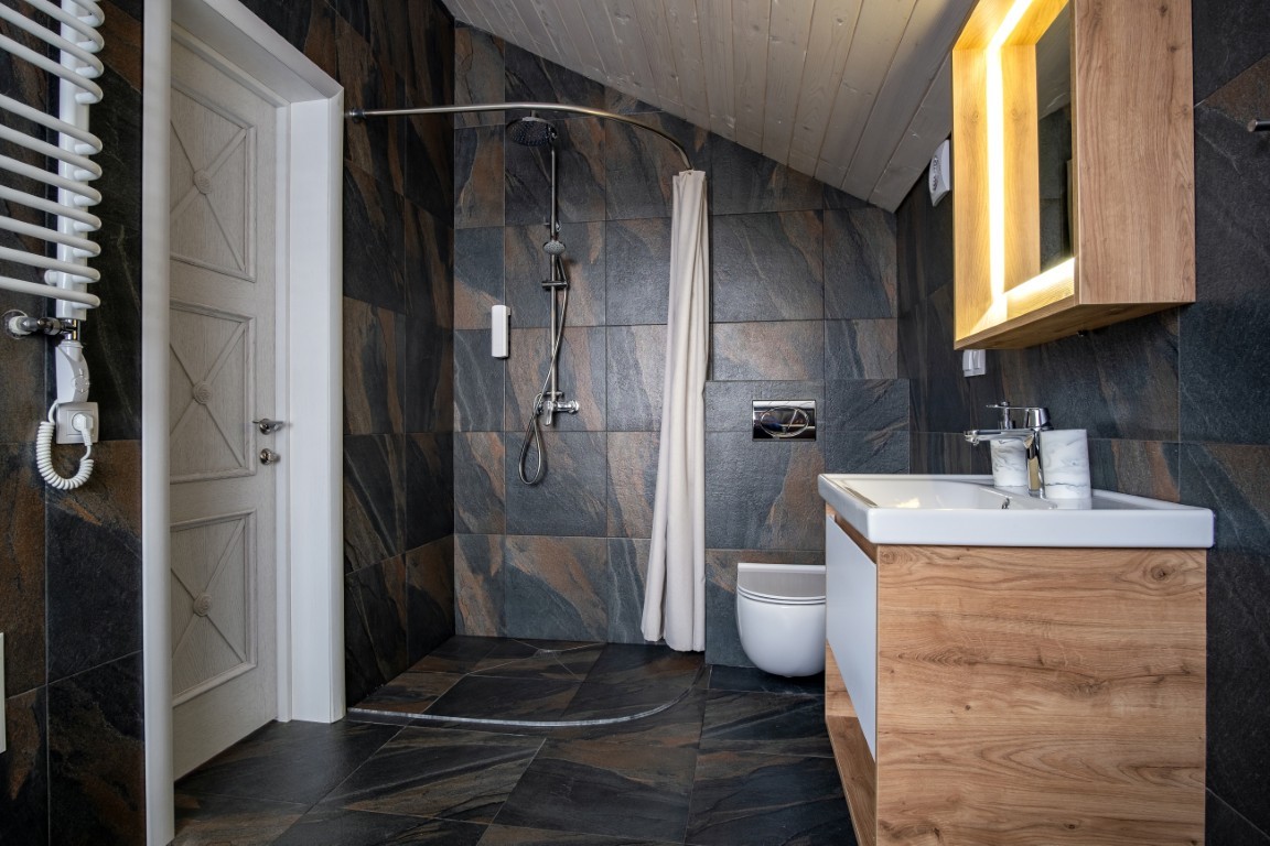 Stylowa łazienka na poddaszu z ciemną podłogą i ścianami z płytek kamieniopodobnych. Wyposażona w walk-in prysznic z zasłoną oraz drewnianą szafkę z podświetlanym lustrem. Połączenie drewna z ciemnymi płytkami nadaje wnętrzu ciepły, luksusowy charakter.
